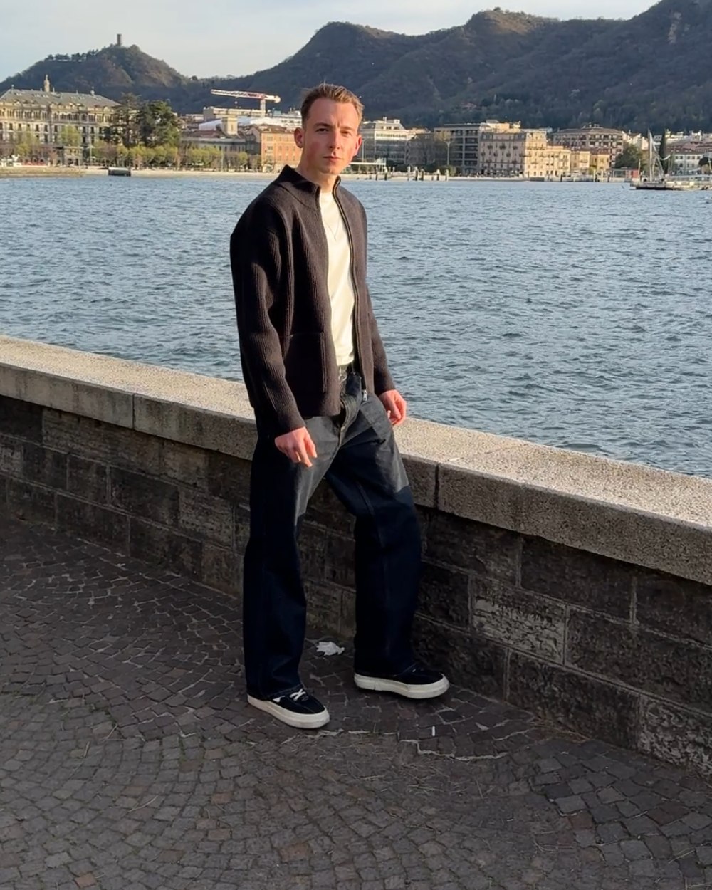 Robin Heinicke an der Uferpromenade des Comer Sees, in Strickjacke, entspannt an eine Mauer gelehnt