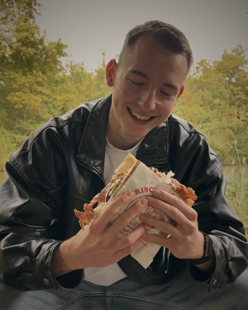 Robin Heinicke sitzt draußen in Lederjacke, hält einen Döner in beiden Händen und lacht